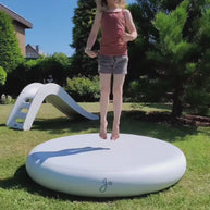 Jong kind springt op witte pad in zonnige tuin met glijbaan op achtergrond.