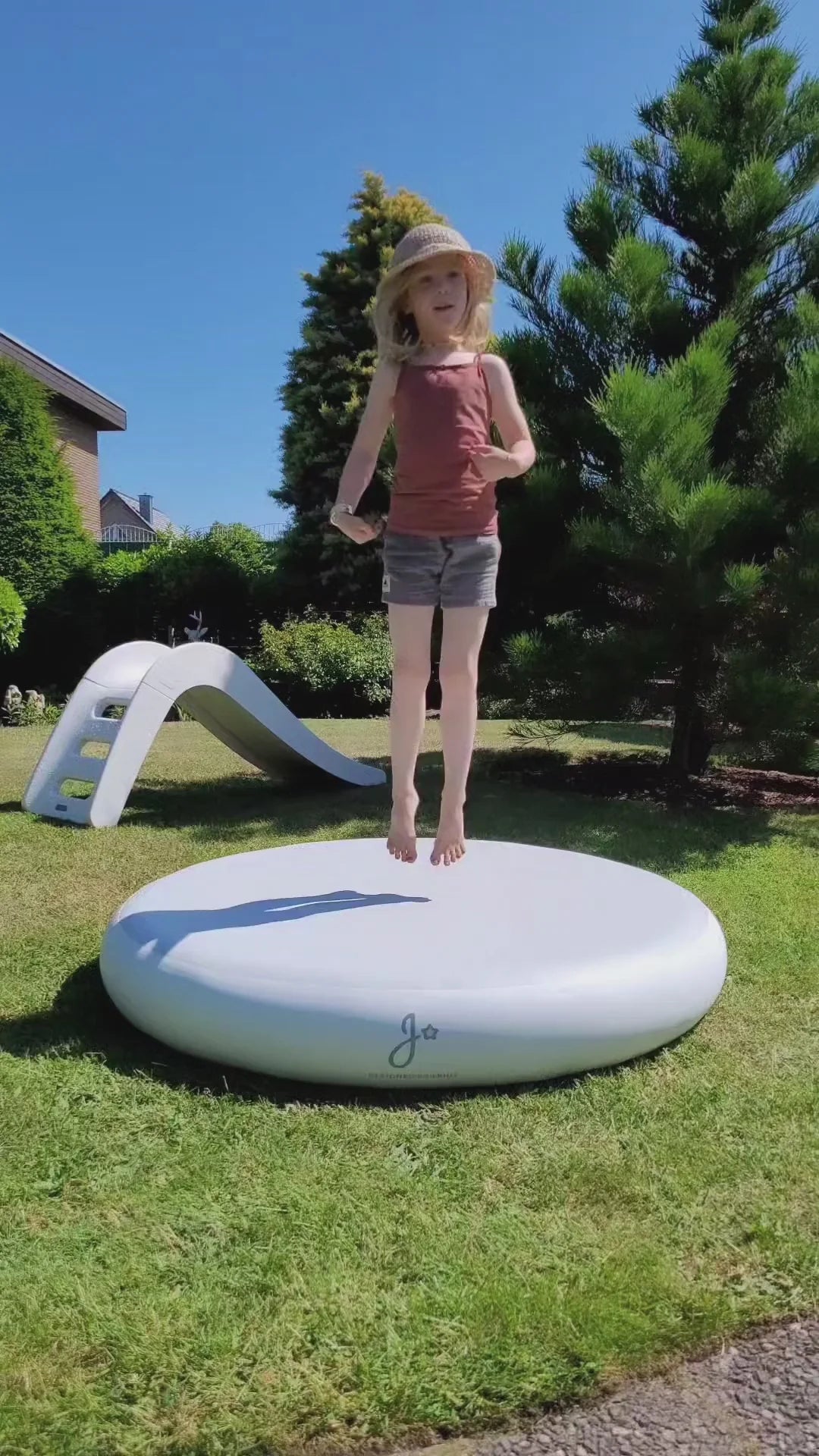 Jong kind springt op witte pad in zonnige tuin met glijbaan op achtergrond.