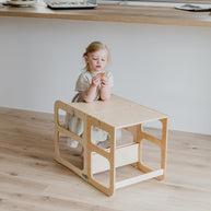 Een jong kind in een lichte jurk zit aan een kleine houten activiteitentafel op een lichte houten vloer, houdt een voorwerp vast en kijkt naar beneden, terwijl de Montessori Houten Learning Tower 2in1 - Hout vlakbij staat en zelfstandig onderzoek aanmoedigt.