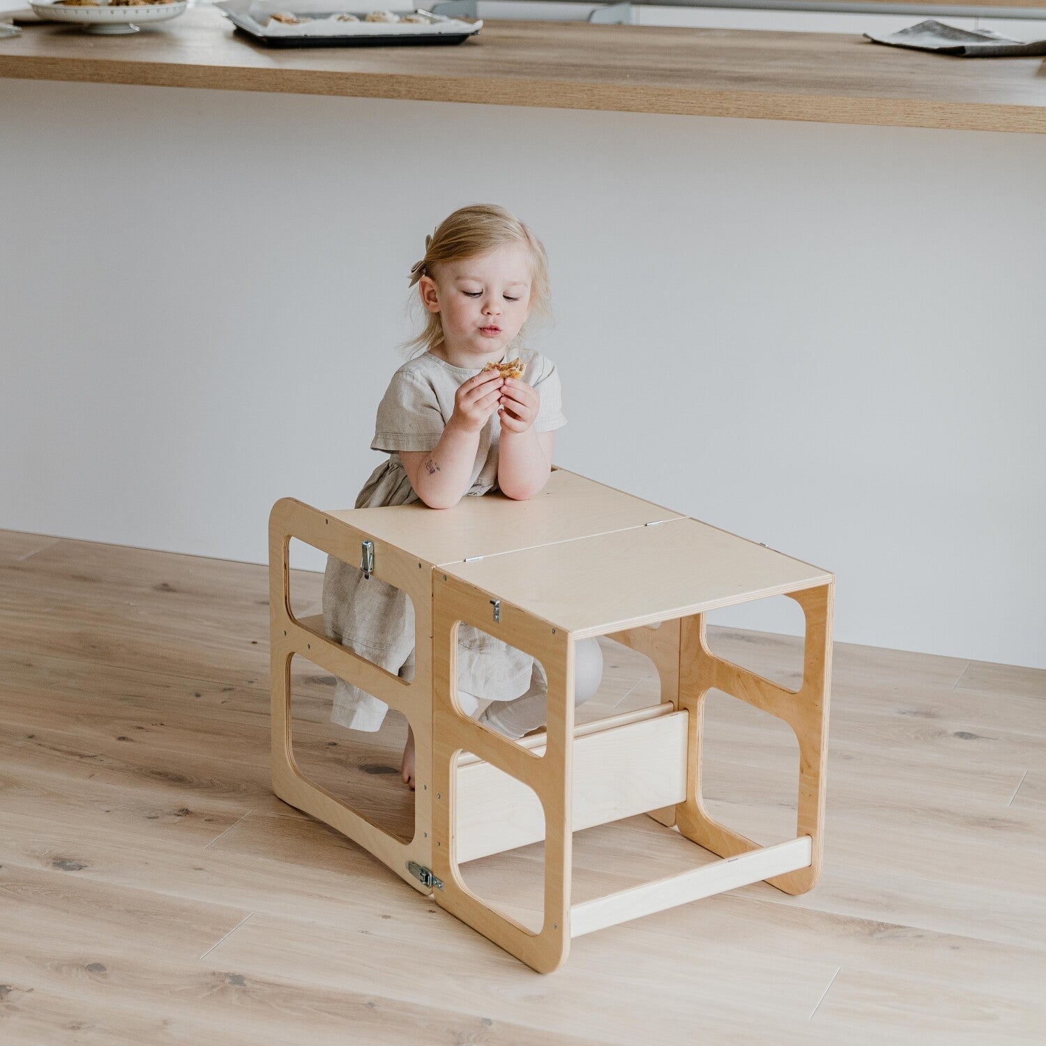 Een jong kind in een lichte jurk zit aan een kleine houten activiteitentafel op een lichte houten vloer, houdt een voorwerp vast en kijkt naar beneden, terwijl de Montessori Houten Learning Tower 2in1 - Hout vlakbij staat en zelfstandig onderzoek aanmoedigt.