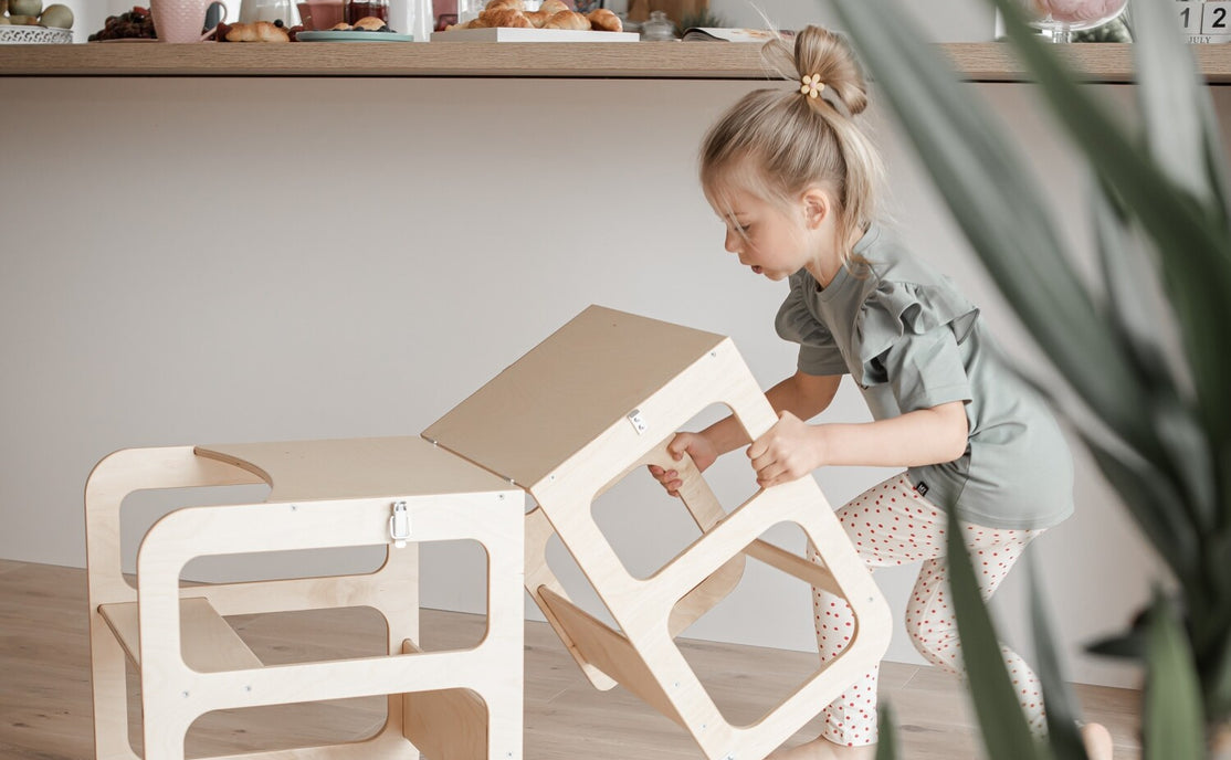 Een jong kind stapelt twee Montessori Houten Learning Tower 2in1 krukjes in een moderne keuken met ontbijtspullen op het aanrecht op de achtergrond.