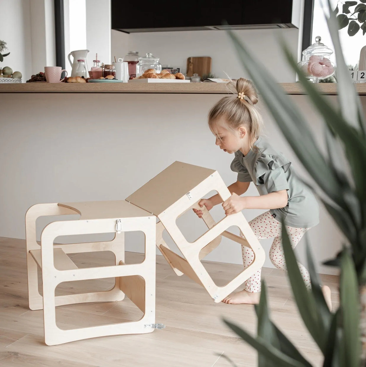 Een jong kind stapelt twee Montessori Houten Learning Tower 2in1 krukjes in een moderne keuken met ontbijtspullen op het aanrecht op de achtergrond.