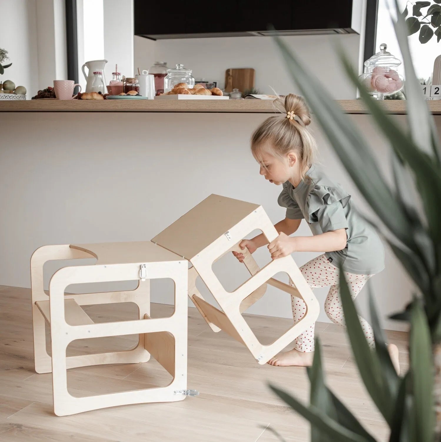 Een jong kind stapelt twee Montessori Houten Learning Tower 2in1 krukjes in een moderne keuken met ontbijtspullen op het aanrecht op de achtergrond.