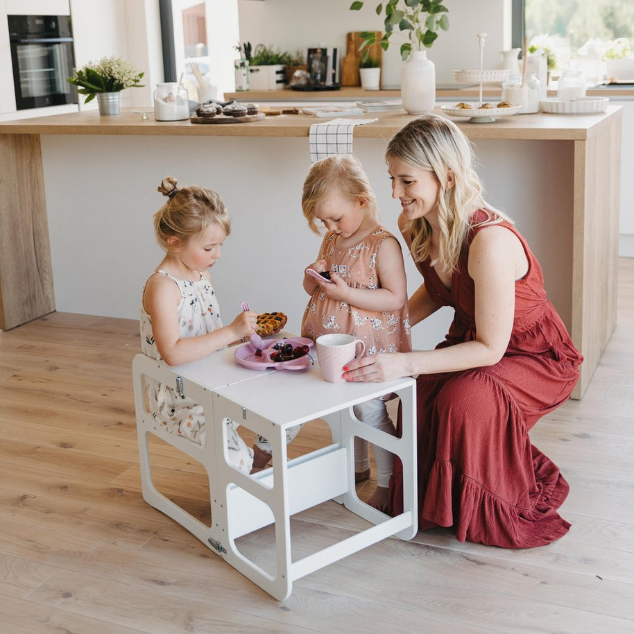 Een vrouw in een rode jurk zit met twee jonge meisjes aan een klein wit tafeltje te genieten van hapjes in een lichte, moderne keuken met de Montessori Houten Learning Tower 2in1 - Wit voor een kindvriendelijke sfeer.