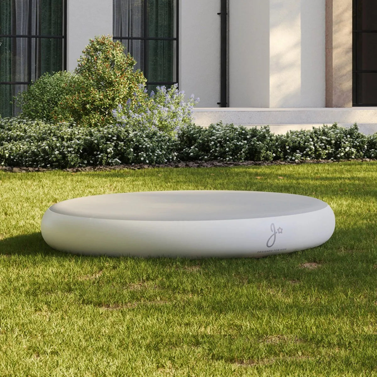 Ronde witte jumpspot (springkussen) op grasveld voor modern huis met groene struiken.
