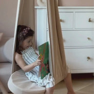 Meisje leest boek in hangstoel, lichte kamer met witte ladekast.