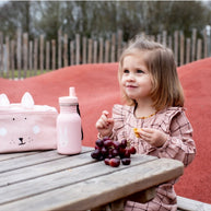 Meisje zit buiten met Trixie Drinkfles Mrs. Bunny 350 ml en fruit op houten tafel