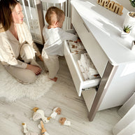Moeder en baby bij de open ladekast naast het cappuccino-witte babybed