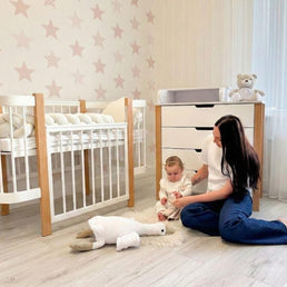 Moeder speelt met baby naast het naturel-witte babybed en commode in de babykamer