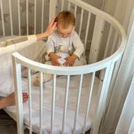 Baby zit in het ronde babybed OLIN met een knuffel en nachtlampje